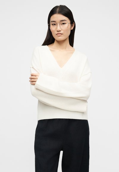 Γυναίκα με μακριά σκούρα μαλλιά και γυαλιά, φορώντας λευκό πουλόβερ με V λαιμόκοψη και μαύρο παντελόνι, στέκεται με σταυρωμένα τα χέρια σε απλό φόντο.