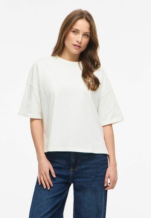 Ung kvinde med brunt hår iført en løs hvid kortærmet skjorte og blå jeans mod en ensfarvet lys baggrund.