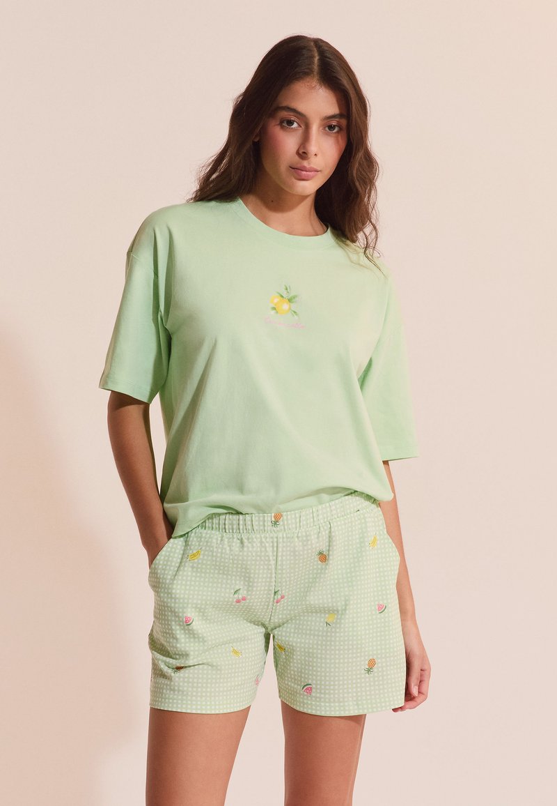 Mujer joven con camiseta de gran tamaño verde claro con gráfico de limón y pantalones cortos verde claro con pequeñas estampas de frutas, de pie con las manos en los bolsillos.