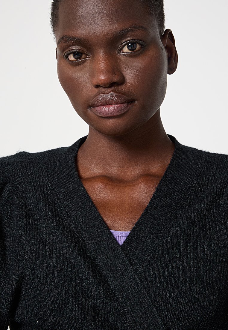 Zwarte gebreide cardigan met een V-hals ontwerp, uitgevoerd in een gestructureerd materiaal met subtiele ribbels. Gekozen boven een paarse kledingstuk.