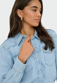 Jasnoniebieska koszula denimowa z wyraźnym kołnierzykiem, zatrzaskami i dwoma kieszeniami na piersi, o luźnym kroju i miękkiej teksturze.