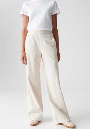 Mujer vestida con camisa blanca de manga corta, pantalones de pana crema de cintura alta y pierna ancha, y zapatillas beige, de pie frente a un fondo liso.