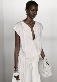White short-sleeve top with a deep V-neck, paired with a flowing white skirt. A textured woven white bag and silver bangles complete the look.