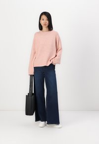 Un sweater oversize rose clair avec des poignets côtelés, associé à un jean bleu foncé à jambe large. Un tote bag noir et des baskets blanches complètent la tenue.