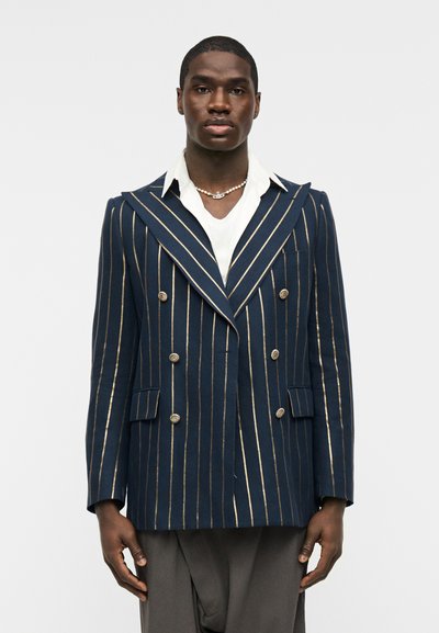 Άνδρας που φοράει ναυτικό διπλόμπρεστο σακάκι με χρυσές κάθετες ρίγες, λευκό πουκάμισο με γιακά, γκρι παντελόνι και κολιέ με μαργαριτάρια, στέκεται μπροστά σε λευκό φόντο.