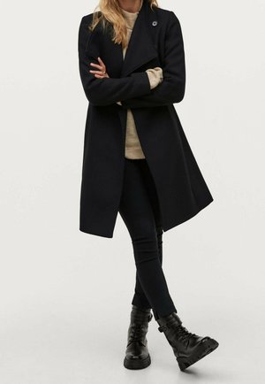 Personne portant un long manteau noir, un pull beige, un pantalon noir et des bottes noires à lacets, debout avec les bras croisés et une jambe croisée sur l'autre.