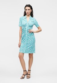 Γυναίκα φοράει τούρκουαζ κοντομάνικο φόρεμα μέχρι το γόνατο με γεωμετρικό σχέδιο, συνδυασμένο με μαύρα ψηλοτάκουνα σανδάλια με λουράκια.