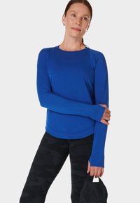 Langemouwen blauwe sporttop met een ronde zoom, zachte textuur en duimgaten bij de manchetten, gecombineerd met zwarte camo leggings en een zwarte pet.