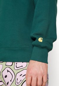 Suéter de algodón verde con puños acanalados; presenta un pequeño logo dorado en la manga. Combinado con pantalones verdes claros de patrón.