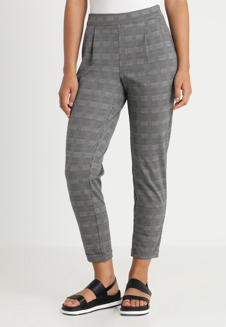 Šedé kostkované kalhoty s elegantním střihem, vysokým pasem a předními záhyby. Materiál má hladkou texturu. Nošeny s černými sandály.