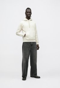 Ανοιχτό μπεζ φούτερ με κουκούλα, μπροστινή τσέπη και ανάγλυφο λογότυπο, σε συνδυασμό με φαρδιά μαύρα τζιν παντελόνια και μαύρα παπούτσια.