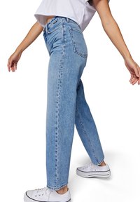 Personne portant un jean droit taille haute bleu clair, un t-shirt court blanc et des baskets blanches, debout les bras légèrement levés.