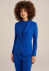 Modrý blazer a odpovídající kalhoty z hladké tkaniny. Blazer má zapínání na jeden knoflík, límec a přední kapsy.
