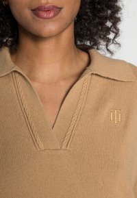 Pull beige en maille avec encolure en V et design texturé. Logo brodé dans une teinte plus claire sur le côté gauche de la poitrine.