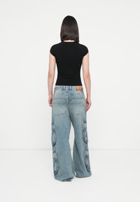 Zwarte aansluitende T-shirt en lichtblauwe wijde cargobroek. Broek heeft grote zijzakken en zichtbare stiksels. Achteraanzicht getoond.