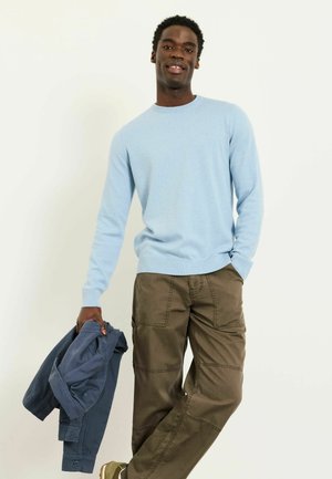 Ein Mann in einem hellblauen Pullover und braunen Hosen steht vor einem schlichten hellen Hintergrund und hält eine gefaltete dunkelblaue Jacke.