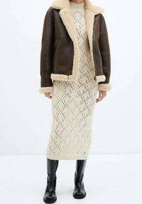 Veste en cuir brun avec doublure en fausse fourrure crème, associée à une robe en tricot crème à motif en losanges et des bottes noires montantes.