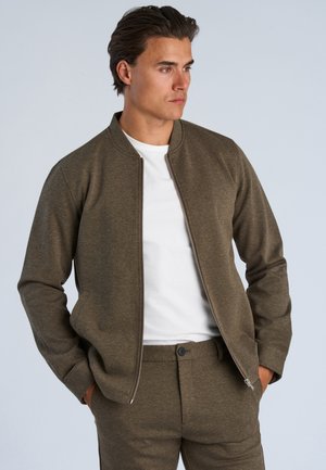 Mand iført olivengrøn lynlåstrøje og matchende bukser med hvid t-shirt, stående med hænderne i lommerne, kiggende til siden.
