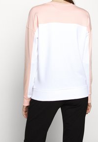 Vrouw in een langemouwen kleurblok sweatshirt met lichtroze en witte panelen, gecombineerd met zwarte broek, gezien vanachter.