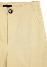 Dettaglio di pantaloni beige che mostra il bottone nero, la tasca laterale, le cuciture e l'etichetta del marchio "Calliope" sulla vita.