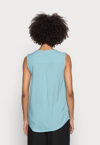 Blouse sans manches bleu clair avec un col rond, présentant un pli discret au dos et un ourlet courbé, confectionnée dans un tissu lisse et léger.