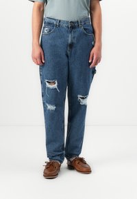 Person wearing blue ripped jeans and brown leather boat shoes, standing against a white background.