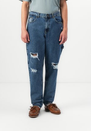 Person wearing blue ripped jeans and brown leather boat shoes, standing against a white background.