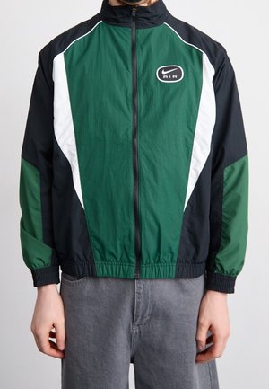 Man wearing green, black, and white Nike Air zip-up jacket with elastic cuffs and gray pants, standing against a plain background.