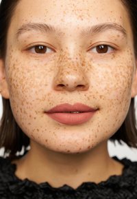 Zbliżenie twarzy o gładkiej cerze z licznymi jasnobrązowymi piegami. Pełne usta pomalowane matową szminką w odcieniu nude.