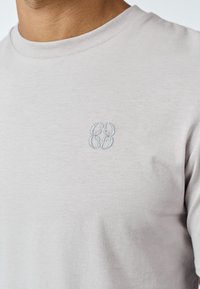 Prim-plan cu o persoană purtând un tricou gri deschis cu un mic design abstract brodat în albastru deschis pe partea stângă a pieptului.