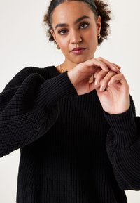 Schwarzer, gestrickter Pullover mit Rippstruktur, weiten Ballonärmeln und lockerem Schnitt. Der Stoff wirkt dick und gemütlich.