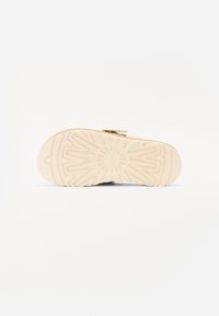 Semelle de sandale avec un motif texturé, de couleur beige et un emblème circulaire au centre. Fabriquée en caoutchouc pour l'adhérence et la durabilité.