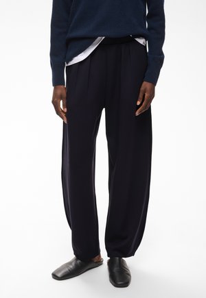 Person trägt dunkle, locker sitzende Hosen, schwarze Slipper, einen dunkelblauen Pullover und ein weißes Hemd, das in der Taille eingesteckt ist.
