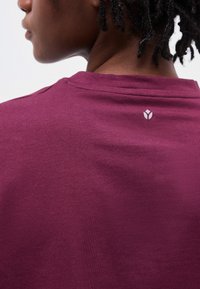 Personne aux cheveux courts et bouclés portant une chemise marron foncé, montrant la nuque arrière avec un petit logo blanc.