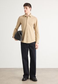 Camisa beige de botón con mangas largas, combinada con pantalones azul oscuro. El modelo sostiene un bolso de piel negra, mostrando un diseño simple y ajustado.