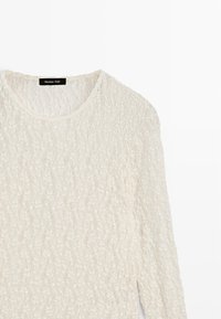 Cream lace long-sleeve top with textured pattern, round neckline, and logo tag at the collar. Fabric appears lightweight and delicate.