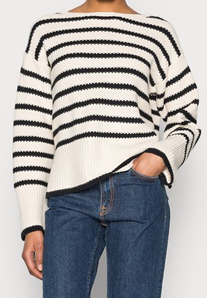 Frau trägt einen weißen Strickpullover mit schwarzen horizontalen Streifen und blaue Jeans, mit einer Hand in der Jeans-Tasche.