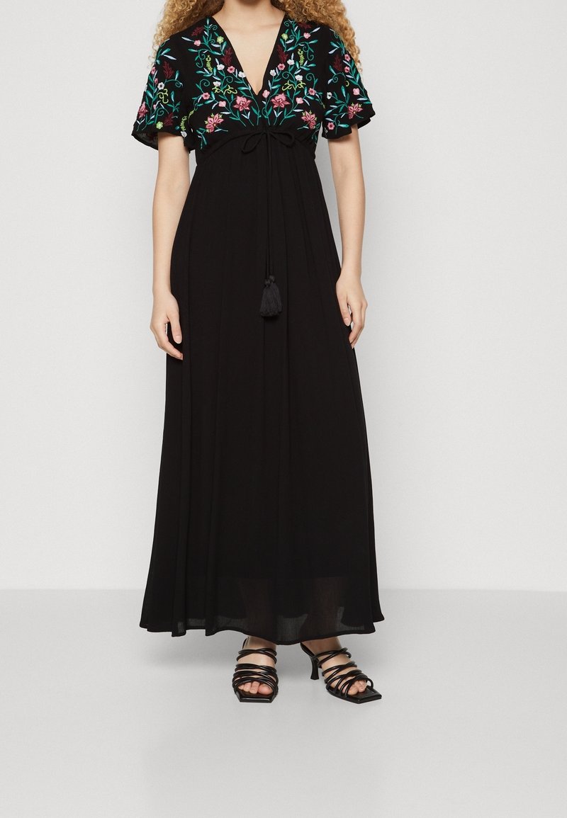 Femme portant une longue robe noire avec des manches courtes brodées de fleurs et des sandales à talons noires à lanières.