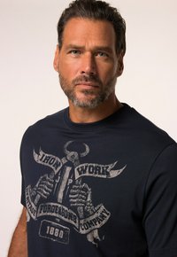 Camiseta de algodón azul oscuro con un estampado gráfico desgastado que muestra manos de esqueleto y texto, incluyendo "IRON WORK" y "1880", centrados en el frente.