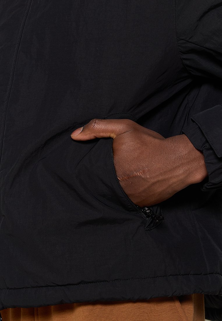 Chaqueta cortavientos negra con un bolsillo con cremallera, puños elásticos y una textura de tela suave y ligera. Detalle de la mano en el bolsillo.