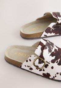 Paire de sandales slides ouvertes à l’arrière avec des lanières imprimées vache marron et blanche et des boucles en laiton réglables sur semelles en liège.