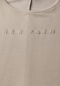 Beige stoffen tanktop met een ronde halslijn. Voorzien van verhoogde, reliëftekst "NEW·PATH." Gladde textuur, minimalistisch ontwerp.