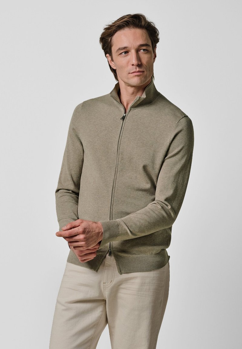 Jasnozielony sweter na suwak wykonany z miękkiego materiału o dzianinowej fakturze, z kołnierzem stójką i ściągaczem u dołu, w zestawieniu z jasnymi spodniami.