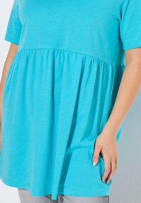 Blusa tunica turchese con maniche corte, vita arricciata e texture morbida; presenta una vestibilità ampia e tasche laterali.