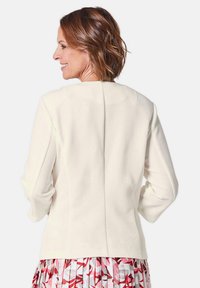 Witte textuurjacket met lange mouwen, voorzien van een naaddetail op de rug, gedragen over een meerkleurige, gemusterde rok.