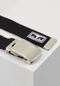 Cinturón negro trenzado con una hebilla metálica plateada y una pequeña etiqueta de marca con un diseño en blanco y negro.