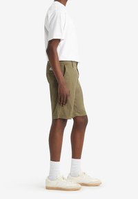 Shorts en coton kaki avec une coupe slim, des poches latérales et une poche arrière avec une étiquette de marque, associés à un t-shirt blanc et des baskets.