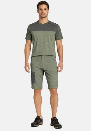 Uomo in piedi che indossa una maglietta verde a maniche corte e pantaloncini verdi abbinati con tasca laterale, scarpe sportive nere, sfondo neutro.