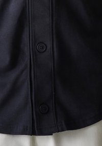 Chemise marine à la texture lisse, avec un plastron à deux boutons noirs et un ourlet droit. Design minimaliste, sans motifs ni accents.