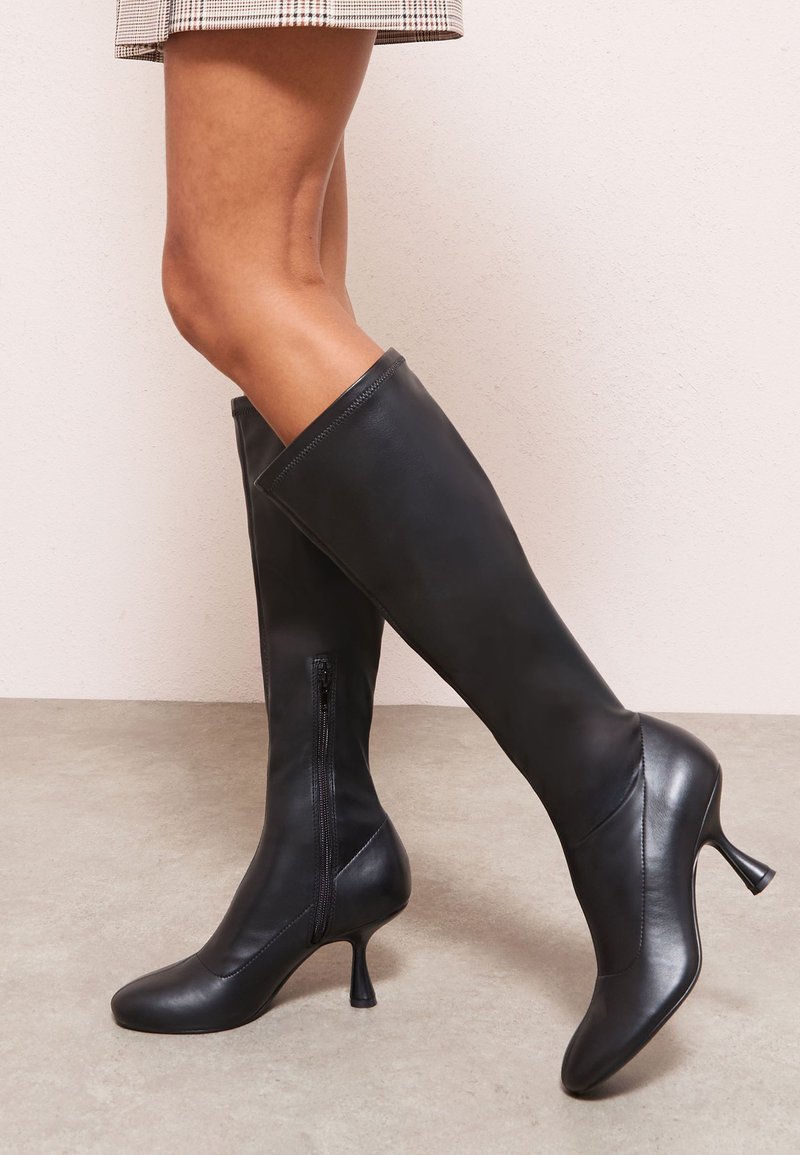 Fekete bőr térdig érő csizma, kúpos kialakítással, hegyes orral és vékony sarokkal. Oldalt cipzárral záródik.